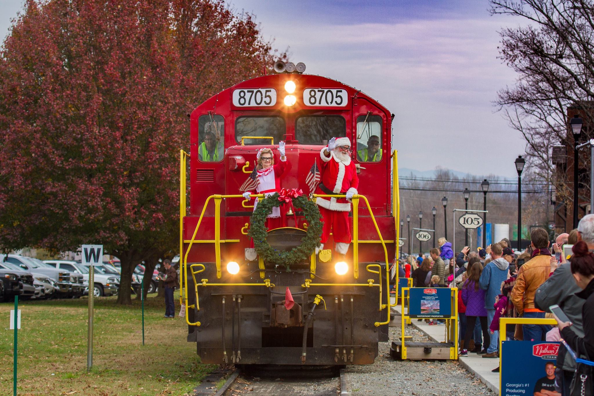 Santa and Mrs. Claus wave to spectators on the Blue Ridge Scenic Railway in Blue Ridge, Georgia. The train is large and red and is adorned with a green wreath at the front of it. Behind the train are rows and rows of people waving back, and a cloudy sky.