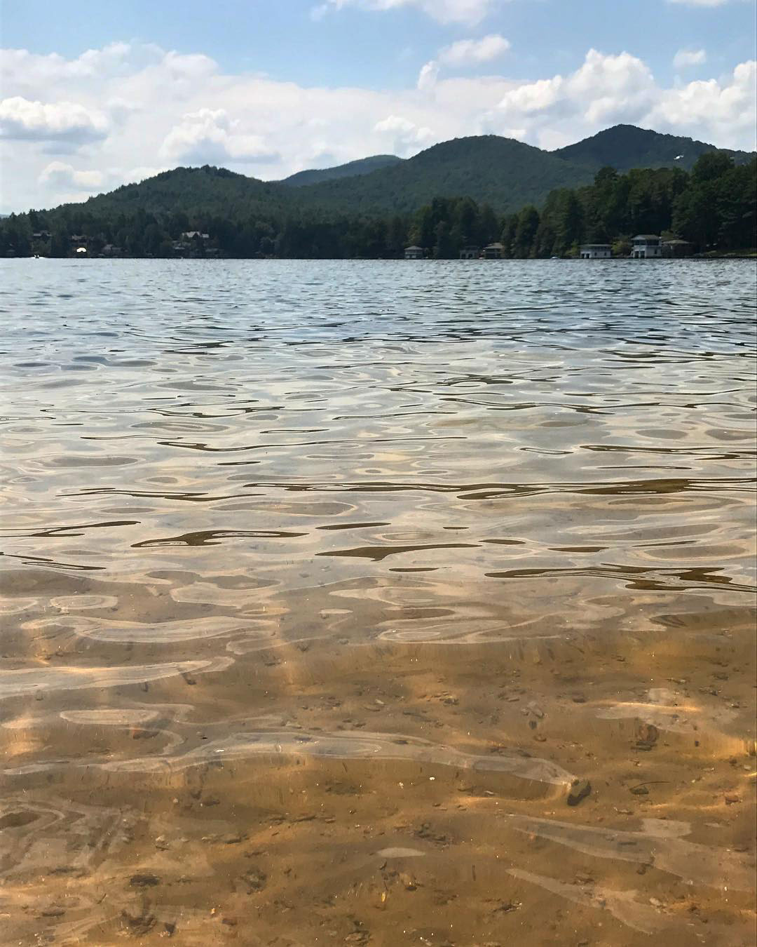 Avertical photo of Lake Rabun Beach in Lakemont, Georgia. The camera is up close with the small waves of the lake, and the water is so clear you can see the brown sand and rocks beneath the surface. In the background are hills of mountains underneath a mostly cloudy sky.
