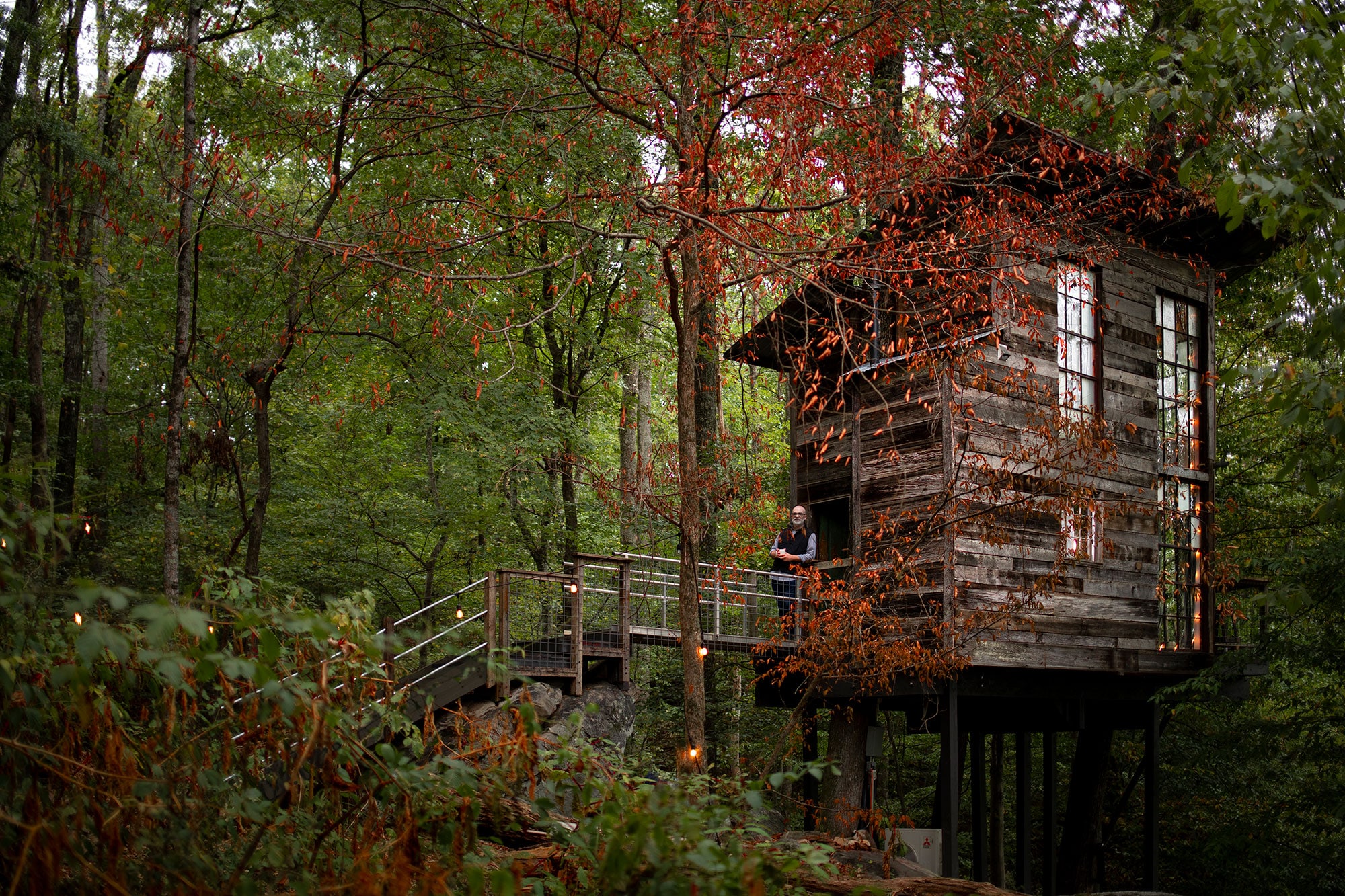 A modern, sleek-looking wooden treehouse stands on stilts amid deep, dense forestry at Treetop Hideaways in Flintstone, Georgia. A person stands on the brideway leading up to the door to the treehouse, leaning on the rails, smiling at the camera. There is a tall, red-leafed tree in front of the cabin, with its leaves just sparse enough to see through them clearly. There are twinkling dangling lights all around the scene.