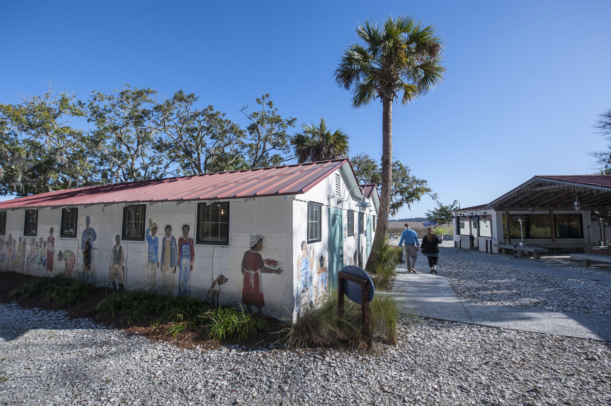 The art-covered, long, red-roofed building of the Pin Point Heritage Museum in Savannah, Georgia, sits on the left of the photo. There is a palm tree stretching up to the sky to the right of the building, and two people next to the building are walking away from the camera. The sky above is deep blue and cloudless.
