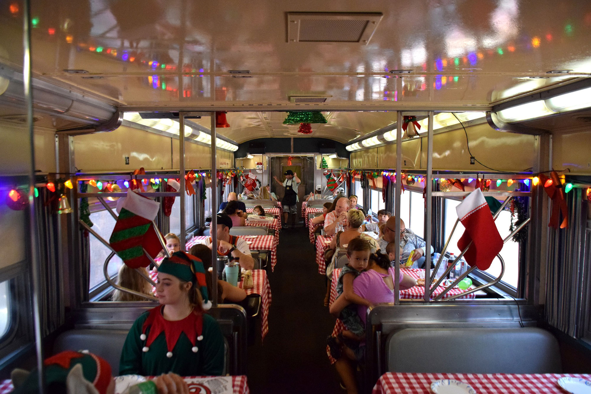 Smiling families and individuals sit in train cars on the Georgia Coastal Railway's Santa Special Train in Kingsland, Georgia. There are red-and-green stockings hanging from the rails and there are also colorful string lights running from one end of the cabin to another.