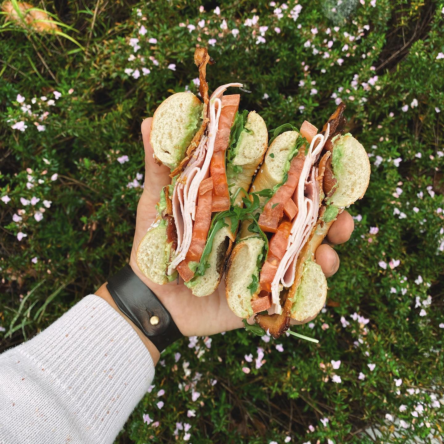 A hand holds two halves of a bagel sandwich with the background of a dark-green shrub littered with bright-pink flowers. The sandwich is from Big Bon Bodega in Savannah, Georgia, and its vibrant colors of ham, bacon, guacamole, arugula and tomato radiate through the photo.