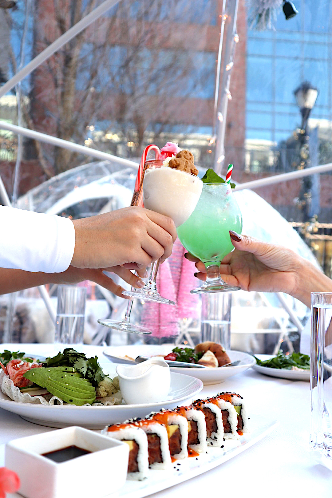 Three hands holding festive drinks over a table of food inside an igloo