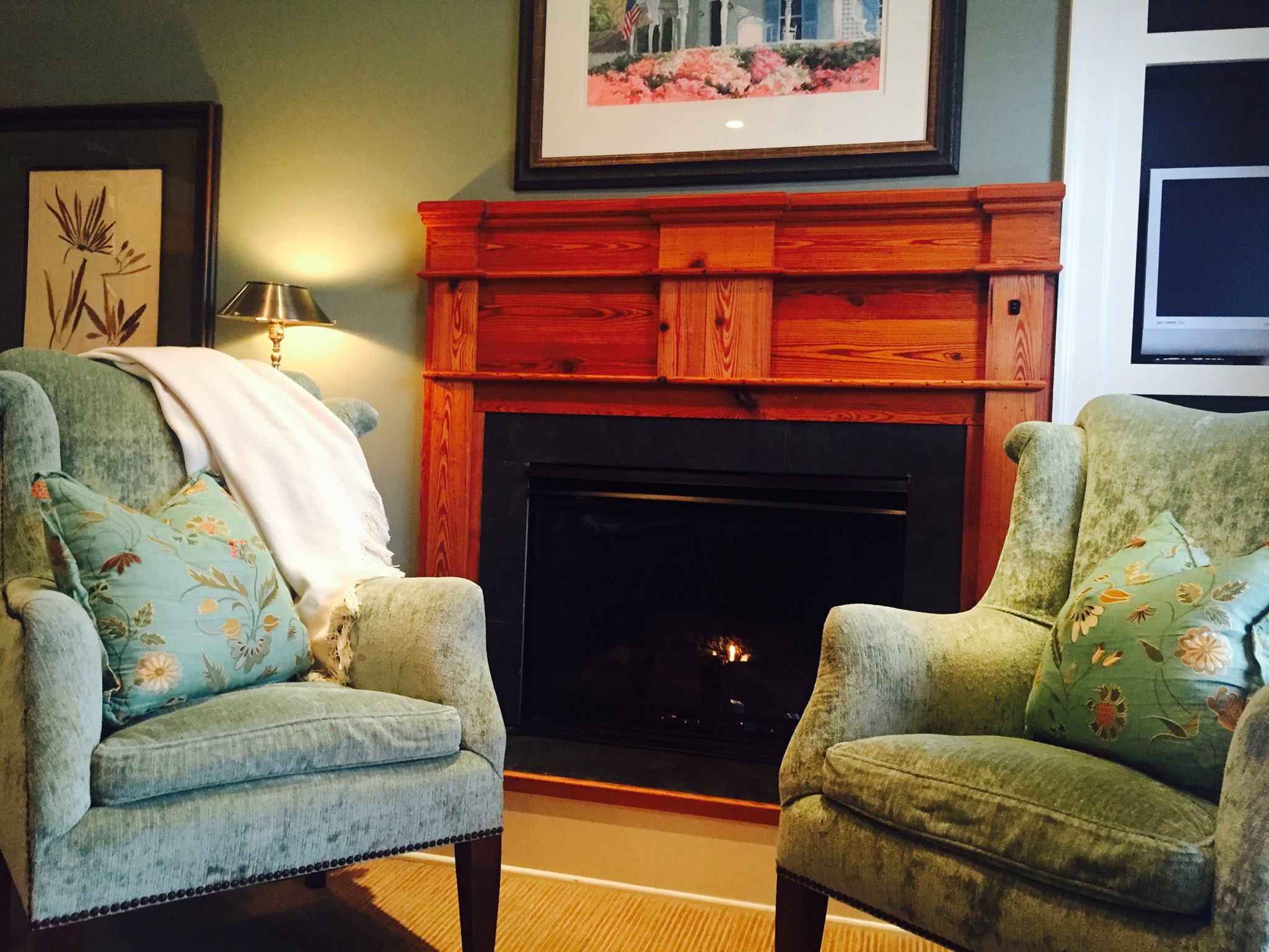 The interior of a cozy room at the James Madison Inn in Madison, Georgia. Two green, velvet arm chairs sit next to each other, with a lit fireplace between them. There are framed pieces of art hung on the walls around the chairs.