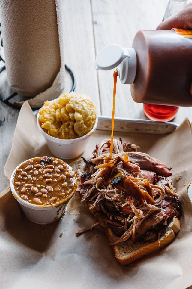 hand pouring barbecue sauce on pulled pork next to containers of baked beans and macaroni and cheese