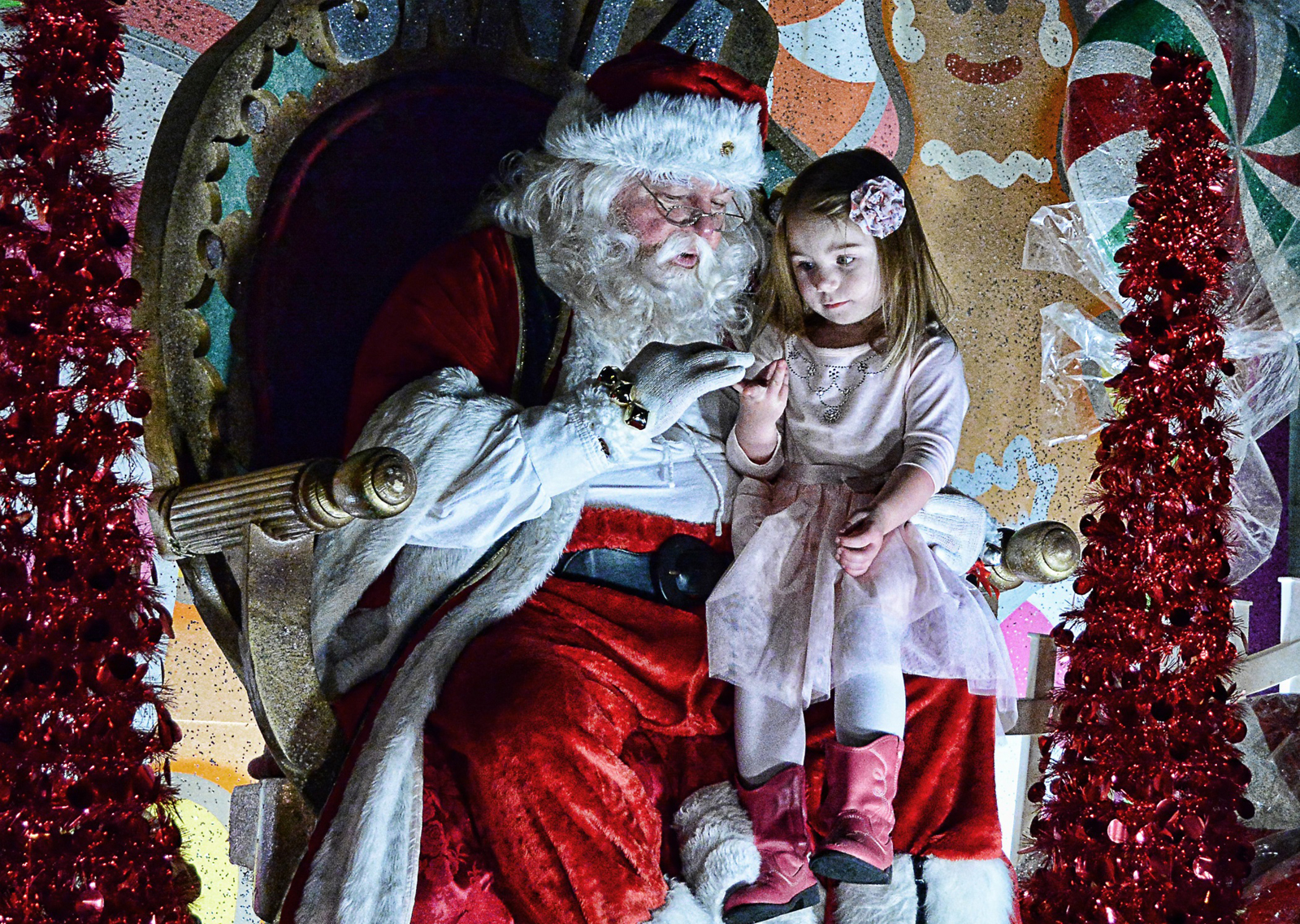 A child wearing red cowboy boots sits on Santa's lap in an extravagant-looking chair at Victorian Christmas in Thomasville, Georgia. The two are holding something and looking down at it in wonder. Santa's suit is very red and their beard is very fluffy and white.
