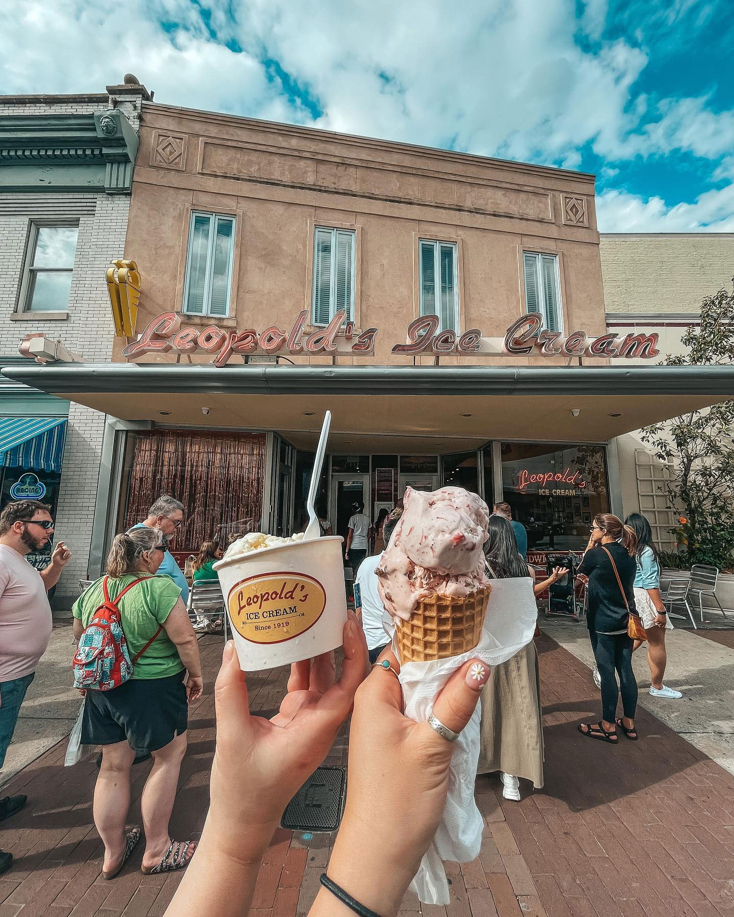 The exterior of Leopold's Ice cream in Savannah, Georgia, is a beige building with two stories and long, vertical windows. In the foreground are two hands holding up a cup of ice cream as well as a waffle cone topped with a few scoops of a pink ice cream. There are more people facing away from the camera in line for ice cream behind the subjects of the photo.