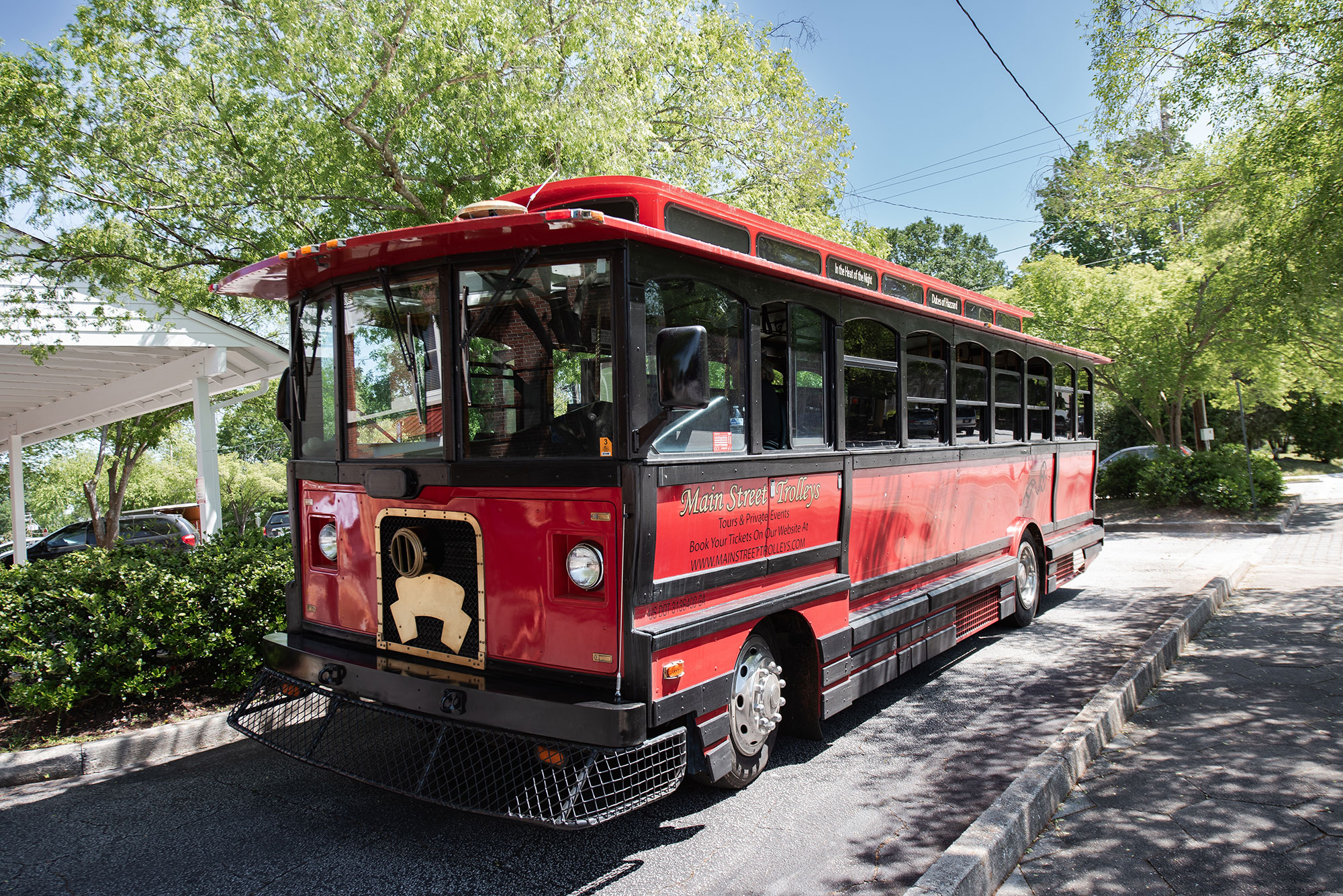 A red-and-black old-school looking trolley, part of the Main Street Trolleys, travels down a road in Covington, Georgia. There are green-leafed trees surrounding the trolley and the sun is shining from a dusty-blue sky.