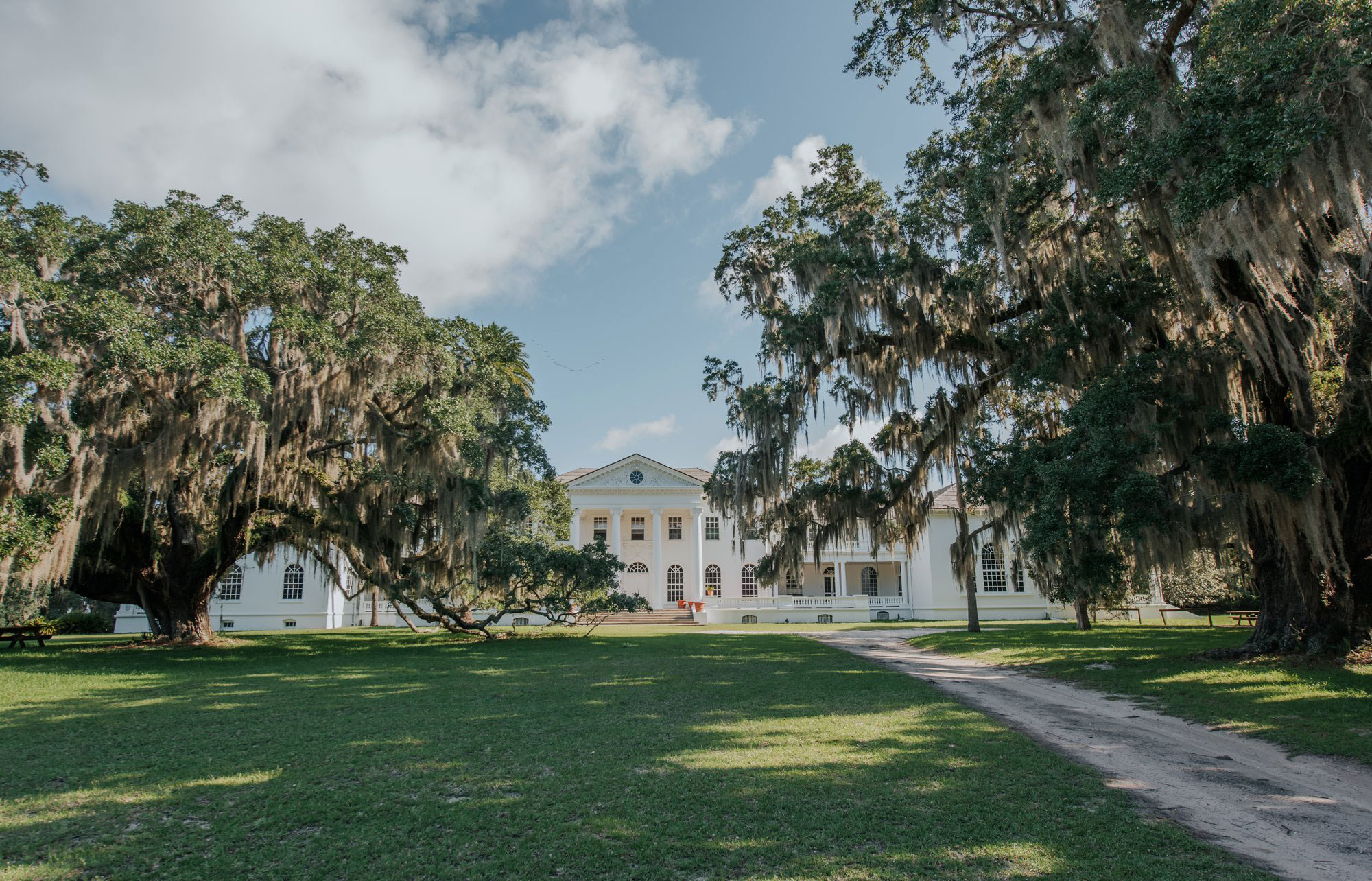 A large, wide, white mansion sits in the middle of the photo under mostly-blue skies. Two willows are on either side of the mansion, which is the Plum Orchard Mansion in Cumberland Island, Georgia. A dirt trail leads to the mansion amid green grass.