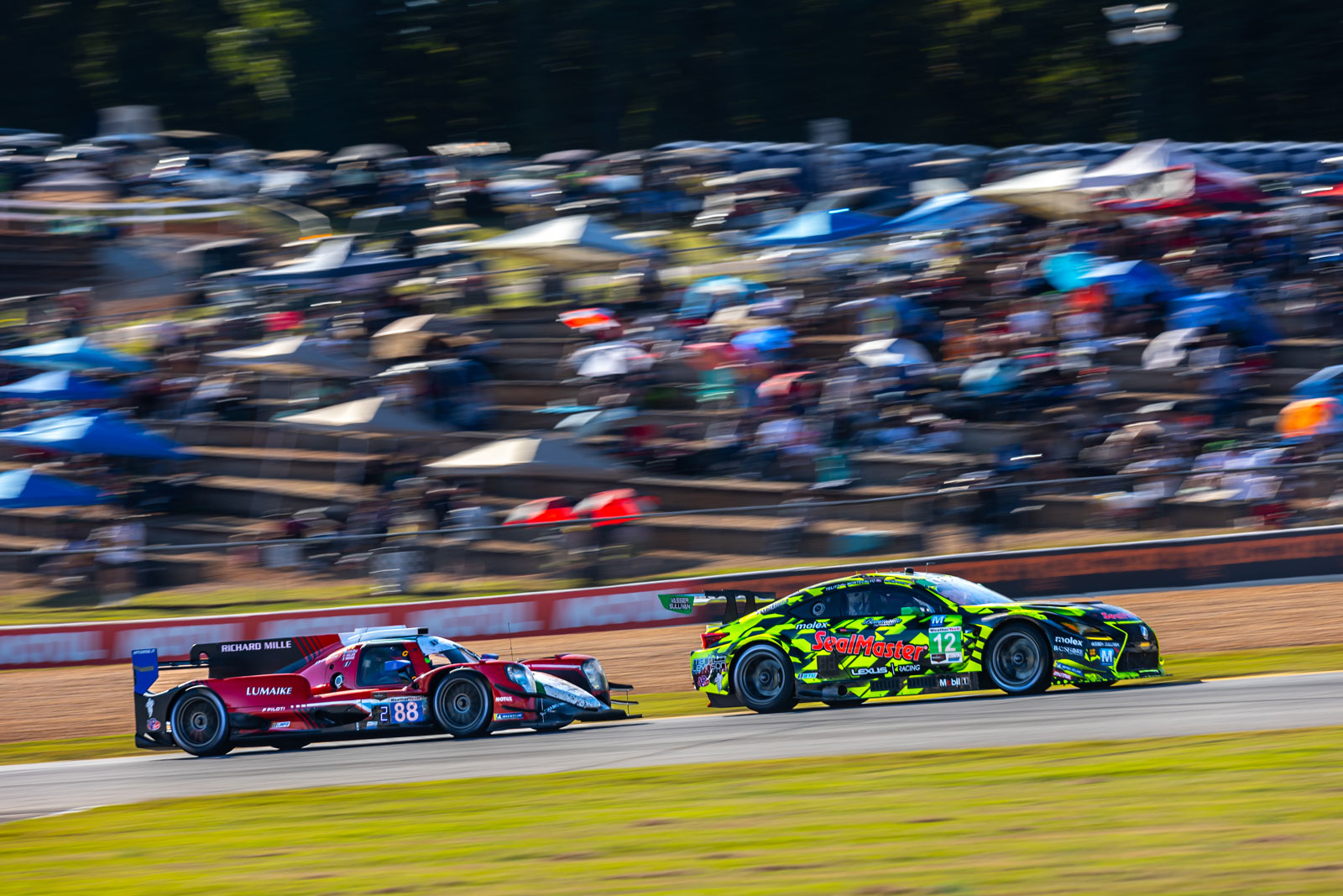 Two race cars zooming with a blurred background of fans and watchers sitting on stands at Michelin Raceway Road Atlanta.