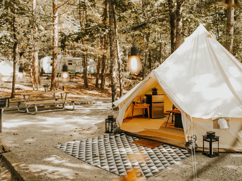 A yurt-looking structure with a cloth roof and walls. Inside, the viewer can see a mini fridge and table with a coffee maker on it. Outside the tent is a large, patterened rug on a dirt square. The surroundings of the glamping tent at Timberline Glmaping Co. in Cumming, Georgia, is scattered tall pine trees and another camper in the distance.