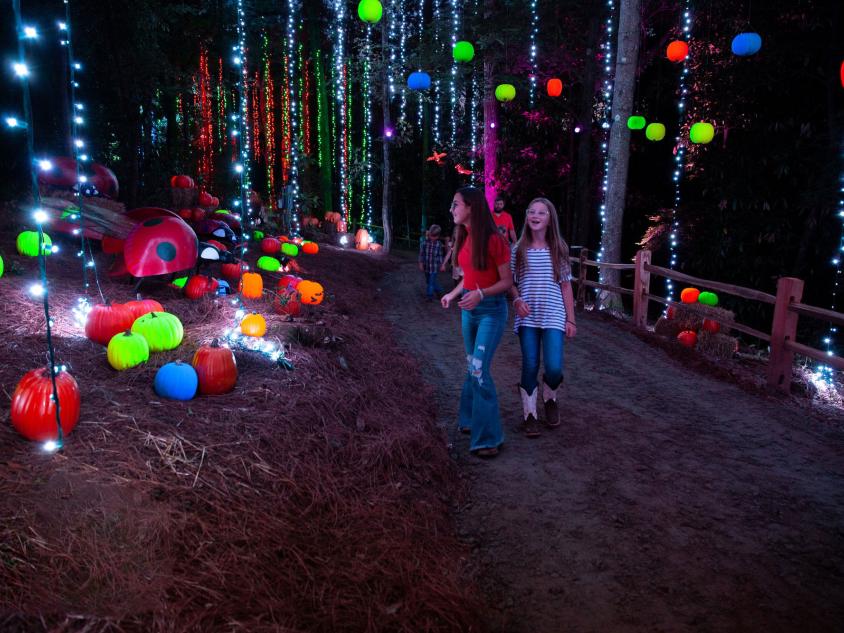 Two teens walking on a trail at night decorated with colorfully lit pumpkins and strings of lights