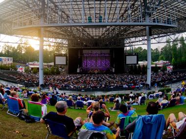 A crowd of concert-goers lying on blankets and fold-up chairs on the green lawn of Ameris Bank Amphitheatre. The amphitheatre is large and metal-ceilinged. In the background is the golden light of the setting sun along with scattered pine trees. On stage is a band getting ready to perform.
