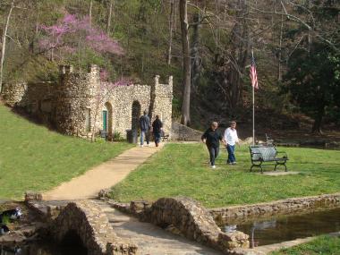 Four people are walking through the green grass and on the dirt path leading up to a stone castle at Rolater Park in Cave Spring, Georgia. There are dense forests behind them and the sun shines on the visitors.