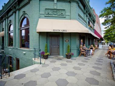 The Mystic Grill storefront in Covington, Georgia. The store's brick exterior is painted a teal blue-green. There are people sitting at the tables outside the store chatting with each other. The sky above is deep blue.