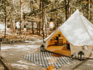 A yurt-looking structure with a cloth roof and walls. Inside, the viewer can see a mini fridge and table with a coffee maker on it. Outside the tent is a large, patterened rug on a dirt square. The surroundings of the glamping tent at Timberline Glmaping Co. in Cumming, Georgia, is scattered tall pine trees and another camper in the distance.