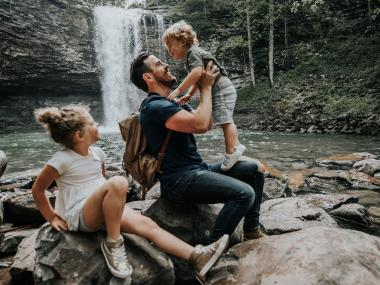 A father and two children splash in the water with a waterfall in the background. 