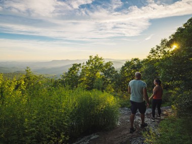 A couple on a hiking trail surrounded by lush greenery and an expansive landscape view in front of them.