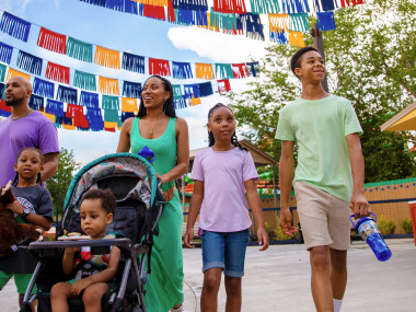 A family walks together beneath colorful banners at an outdoor attraction, with young children, a stroller and a taco stand nearby.