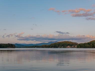 The sweeping Lake Chatuge in Hiawassee, Georgia. There are mountains in the background and forests of dark-green trees. The sky above is pastel blue and pink from sunset.