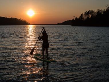 A stand-up paddleboarder stands upright on their blow-up paddleboard, slightly deflated, floating on the gentle waves of Carters Lake in Ellijay, Georgia. They have a paddle in their hands and are actively paddling. In the background are a few shorelines lined with tall trees, and in the sky before the paddleboarder is the setting sun, which paints the entire scene in a golden glow and reflects its shine off the water.