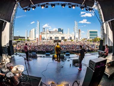 A band plays for a large crowd outdoors with a city skyline in the background.