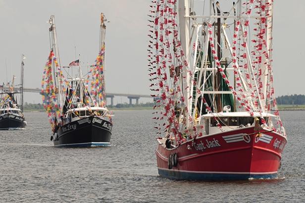 Three ships with decorative flags float along the water as part of the Blessing of the Fleet in Darien, Georgia. The sky above is gray and in the background is a large bridge.