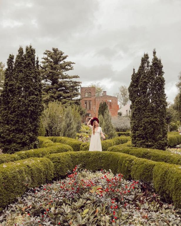 A woman walks through a lush garden with a large house in the background