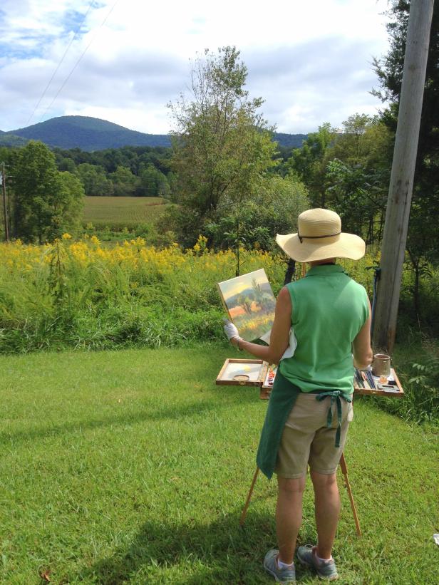 Photo of artist facing away from camera painting a picture outdoors with trees and flowers in the background in Blue Ridge, Georgia