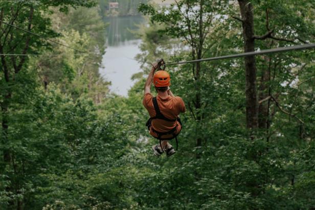 Someone zooms down a zipline at Unicoi State Park in Helen, Georgia, their back to the camera. They are wearing protective gear and flying down the zipline through an opening in a dense, green forest.
