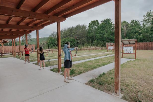 Three people shooting archery under a covered pavilion with targets in the field