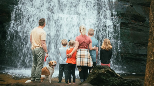A family with four children and a dog facing a tall waterfall with rocky edges.