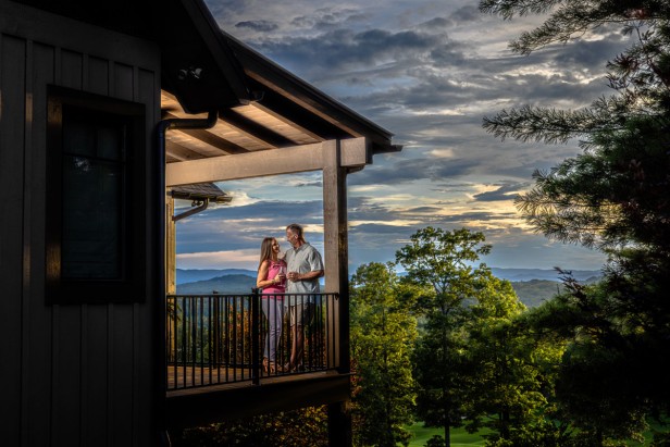 A couple holding wine glasses standing in the corner of a balcony high above near the trees with a cloudy sky above.