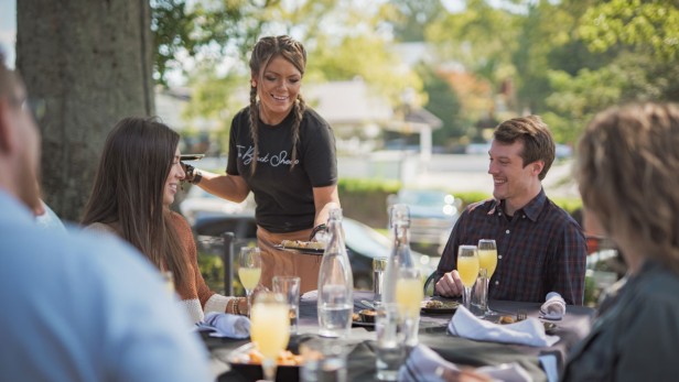 A server with a black shirt serving a table of all adults with mimosas and plates of food taking place outside.