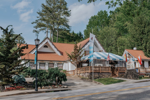 A red-roofed Bavarian-looking building sits on a street in Helen, Georgia. The restaurant is Hofer's of Helen. There are tall trees behind the building under blue skies.