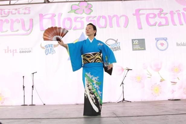 A performer in traditional-looking Japanese robe holding out a fan on a stage at the Conyers Cherry Blossom Festival in Georgia.