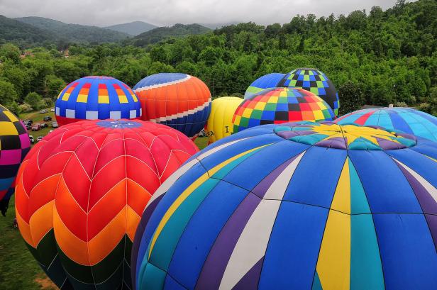 Around 10 hot-air balloons sit inflated on the ground (which is not visible), preparing for the Helen to the Atlantic Balloon Race and Festival in Georgia. The tops of the hot-air balloons are colorful with geometric shapes and multiple colors. Behind the balloons is a dense, dark-green forest of trees, behind which are misty tops of mountains under a gray-cloud sky.