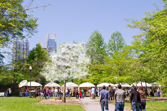 Green-leafed trees line the green lawn of Piedmont Park in Atlanta, Georgia, as part of the Atlanta Dogwood Festival. There are several spectators and white-topped tents. The sky above is clear and blue.