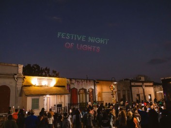 Festive Night of Lights celebration with crowds enjoying illuminated historic buildings and drone light show spelling "Festive Night of Lights" overhead. Join us in Olde Town Conyers Entertainment District.