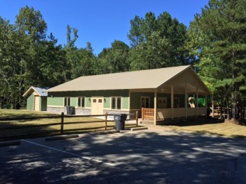 Available for RENT: Retreat House at Black Shoals Park & Randy Poynter Lake in Conyers/Rockdale County, GA. Charming countryside lodge with green shutters and covered porch, nestled among tall pine trees under clear blue skies, ideal for retreats.