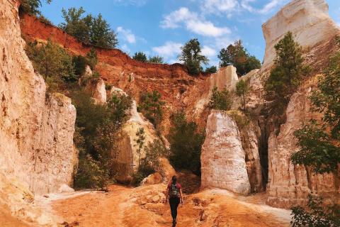 A hiker wearing a backpack faces away from the camera and walks along the rusty orange ground of Providence Canyon State Park in Lumpkin, Georgia. In front of the subject of the photo are the tall sandstone-looking structures jutting out from the ground, littered with dark-green-leaved trees. The sky above is blue with small puffy clouds.