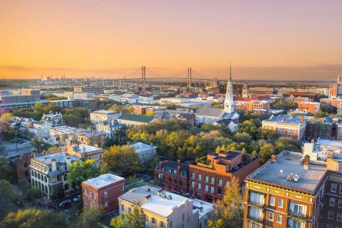 An aerial view of historic Savannah, Georgia. In the foreground are several antique brick buildings surrounded by trees among the streets; the lower half of the photo includes scenery like this, and along the horizon is the Talmadge Memorial Bridge. The sky is painted orange and purple by a Georgia sunset.