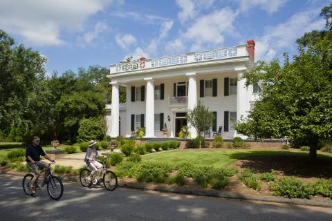 Two people on bikes cycle past a large historic mansion in the background.