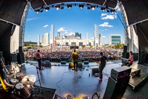 A band plays for a large crowd outdoors with a city skyline in the background.