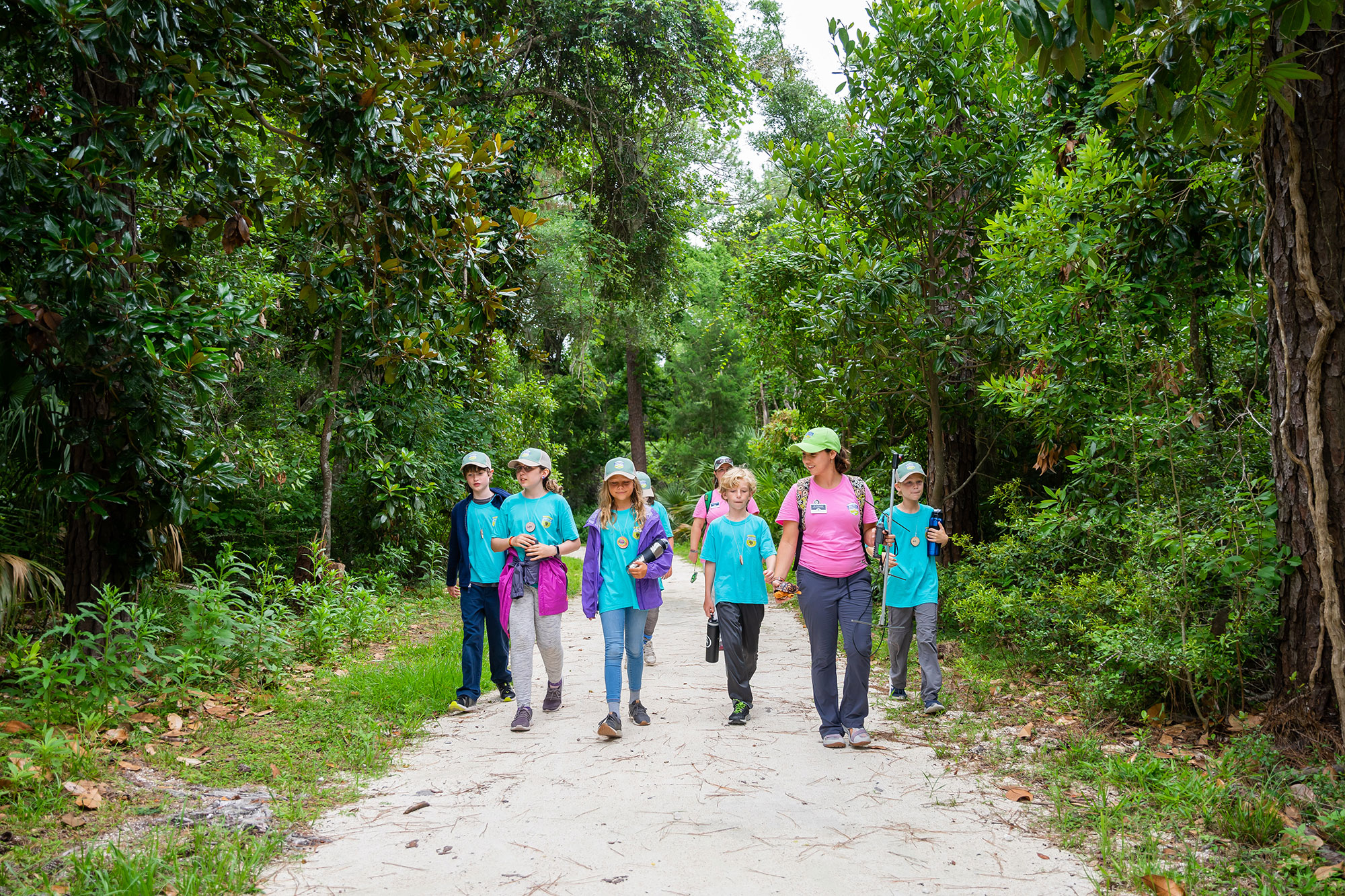 Children in blue shirts walk on a sandy path with adults in pink shirts during summer camp on Jekyll Island, Georgia