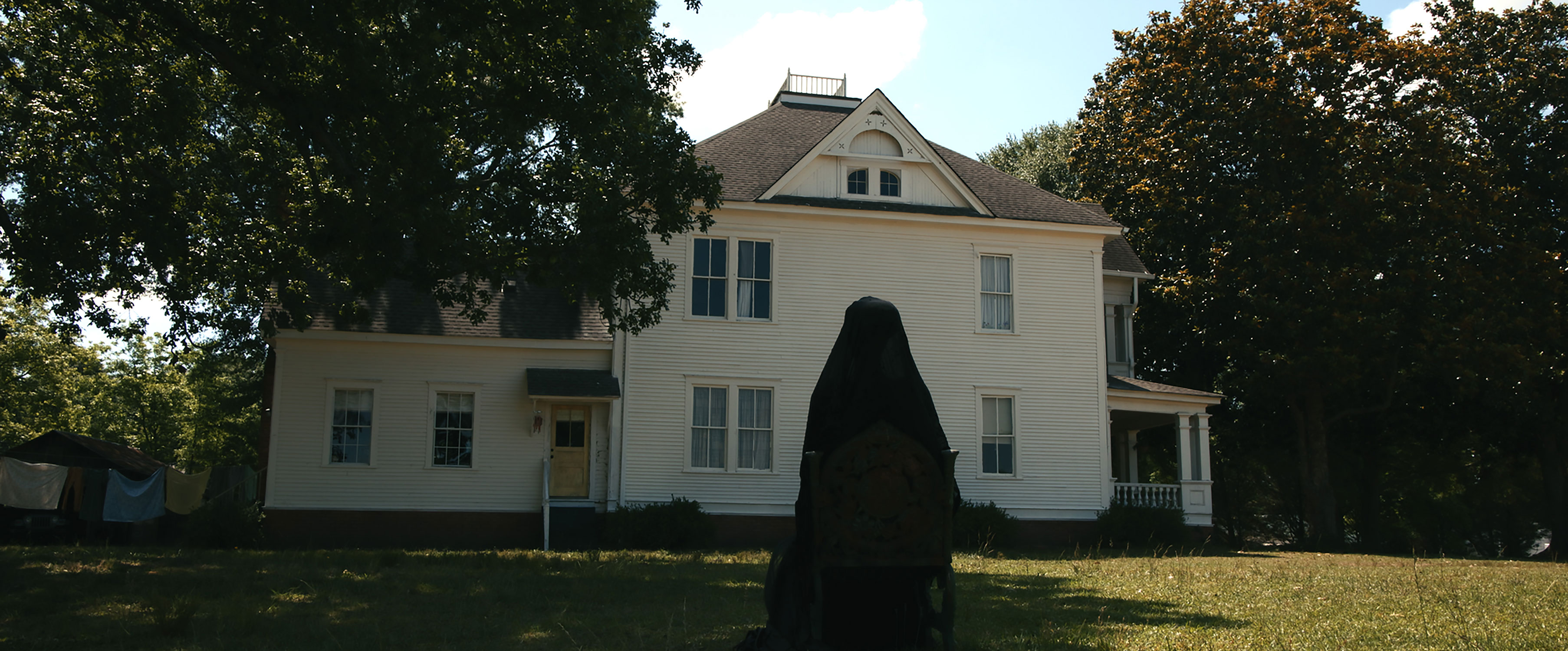 A woman draped in black stands in the yard in front of a white house in a scene for "The Woman in the Yard"