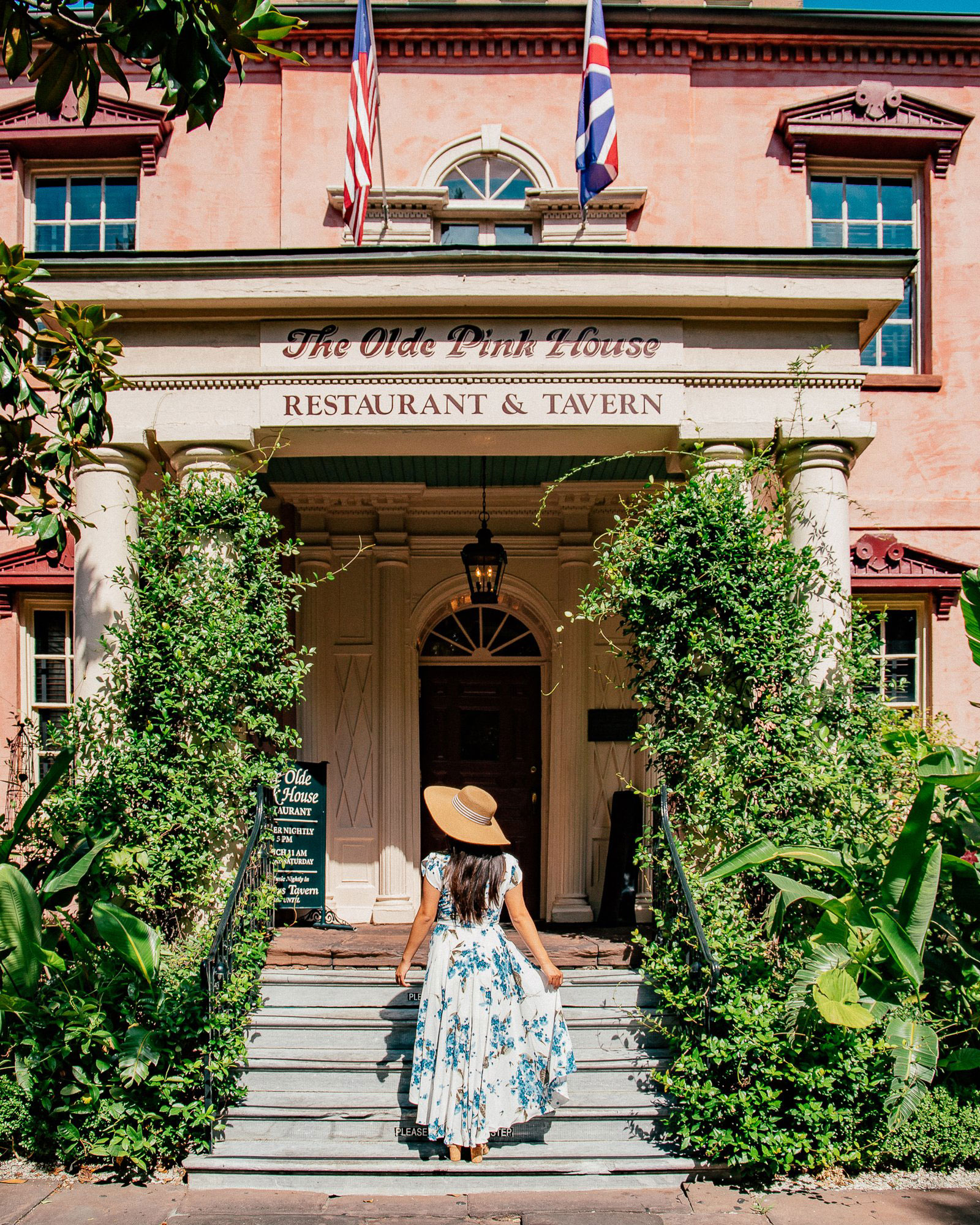 Woman in a straw hat and blue and white floral dress walking up stairs to The Olde Pink House
