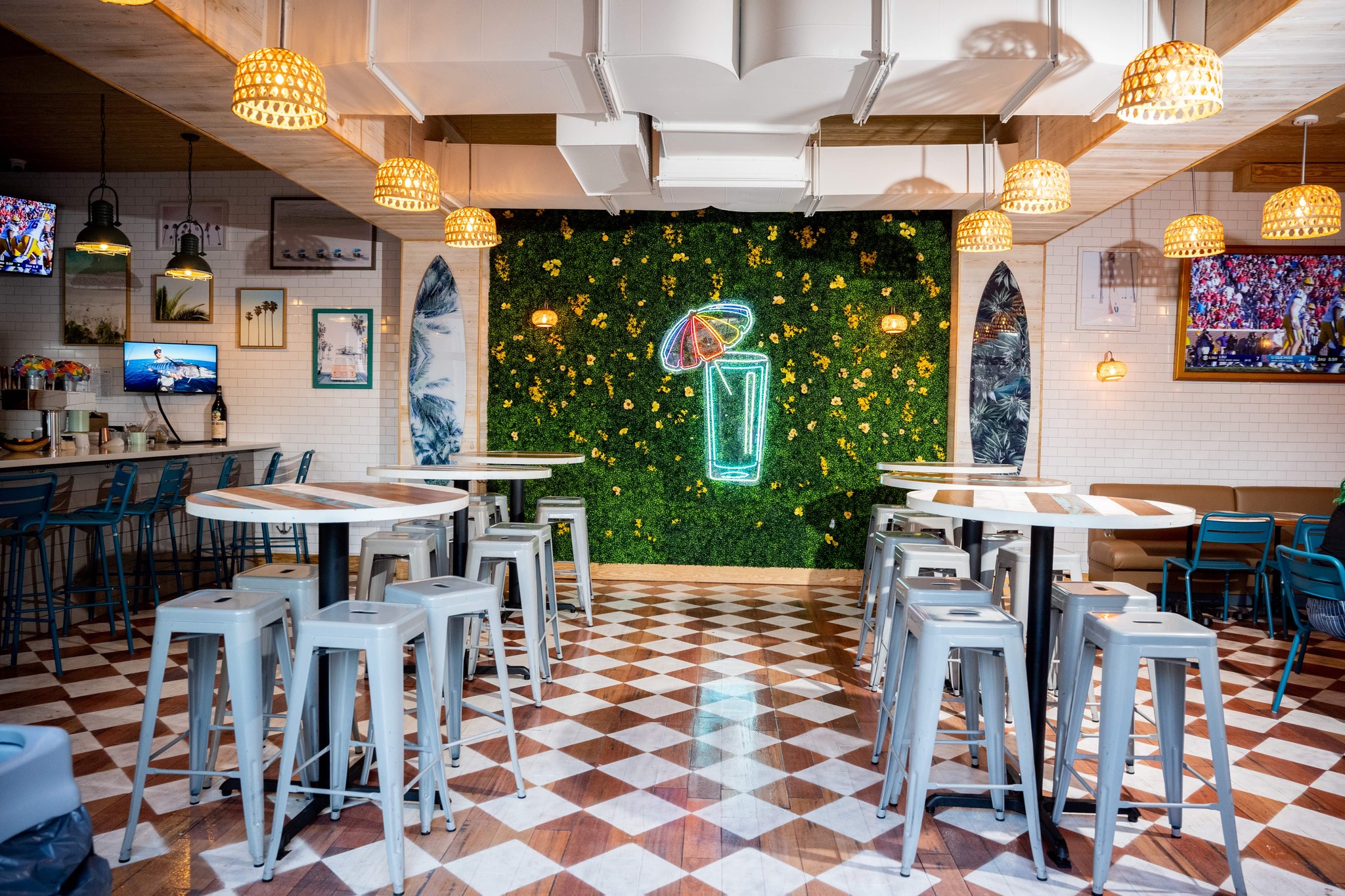 The interior of Zunzibar in Savannah, Georgia, features a lot of colorful and flashy decor, lights and futniture. In the middle background of the phtoo is a grass wall with a neon-light display of a blue glass with a rainbow paper umbrella. There are stools and tables lined up across the room next to other seating, and the floor is decorated in checkerboard pattern.